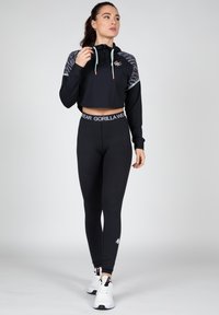 Zwarte cropped sweater met patroonmouwen, capuchon met trekkoord en logo. Zwarte legging met merknaam bij de taille. Witte sportsneakers.