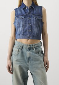 Cropped denim vest in dark blue with sleeveless design, featuring two front pockets and button closure. Paired with light blue, loose-fit jeans.