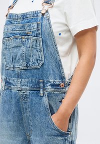 Άτομο που φοράει ανοιχτό μπλε τζιν φορμάκι με μπροστινές τσέπες και λευκό μπλουζάκι με μικρή μπλε καρδιά, χέρι στην πλαϊνή τσέπη, σε γκρι φόντο.