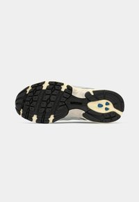 Semelle en caoutchouc noire et beige d'une basket avec un motif texturé et trois petits accents ovales bleus près du centre du talon.