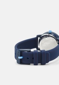 Reloj de silicona azul marino con correa texturizada, acentos azules y hebilla de metal. La caja redonda presenta un fondo reflectante con el logotipo de la marca.