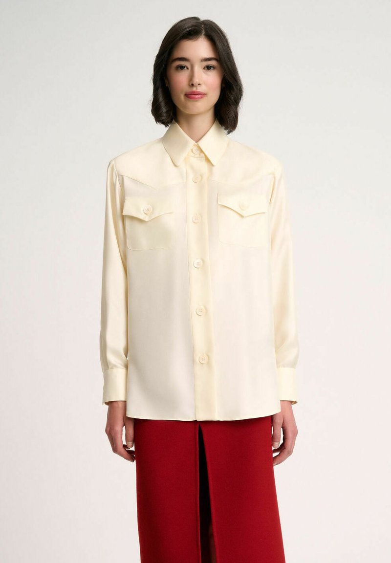 Camicia di seta color crema con colletto a punta, maniche lunghe e due tasche sul petto; presenta una chiusura con bottoni sul davanti e una texture liscia.