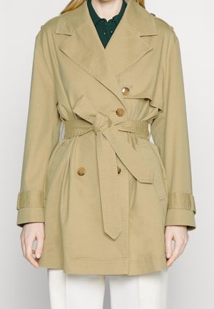 Beige dobbeltradet trenchcoat med knapper og bundet bælte, båret over en mørkegrøn skjorte med krave og hvide bukser.