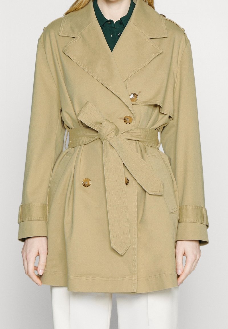 Trench-coat beige croisé avec boutons et ceinture nouée, porté sur une chemise à col vert foncé et un pantalon blanc.