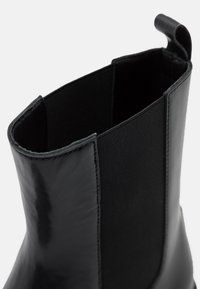 Sorte ankelstøvler med en blank læderoverflade og elastiske sidepaneler. Tekstureret syning langs kanterne tilføjer detaljer.