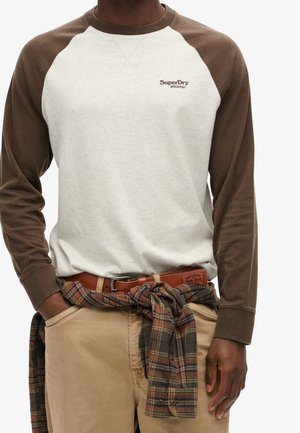 Beige-brauner Raglan-Langarmshirt mit kleinem "SuperDry"-Logo, kombiniert mit beigen Hosen und einem karierten Flanellhemd, das um die Taille gebunden ist.