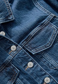 Ciemnoniebieska tkanina dżinsowa z podwójnymi szwami, kieszenią z klapką i metalowymi guzikami z grawerem "NEXT JEANS SINCE 1982."