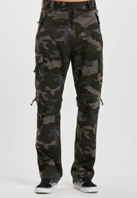 Pantalons cargo décontractés en camouflage vert foncé et noir, avec des poches à zip et à rabat, portés avec des baskets noires et blanches.
