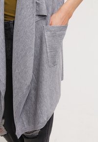 Cárdigan gris hecho de una tela suave, con un frente abierto, acabado texturizado y un bolsillo lateral. Combinado con una blusa verde y jeans negros.