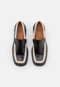 Chaussures à enfiler en cuir noir avec accents beige, design à bout carré et semelle en caoutchouc texturée. Dessus lisse avec détails matériels minimaux.