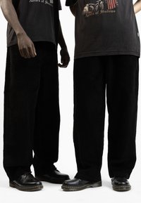 Pantaloni a gamba larga in corduroy nero con tessuto testurizzato, abbinati a t-shirt grafiche nere e scarpe nere con cuciture gialle e suola grossa.