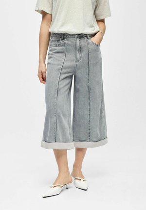 Osoba nosi svetlošedé široké krátke rifle s vyhrnutými lemami, svetlom farebnú košeľu s krátkym rukávom a biele špicaté nízke topánky na podpätku.