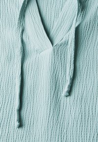 Tessuto blu chiaro con texture e un sottile motivo a pieghe. Presenta una scollatura a V e cordini con piccoli nodi alle estremità.