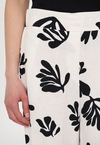 White linen trousers featuring bold black floral patterns, textured fabric, wide waistband, and a side pocket design.