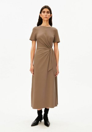 Frau trägt ein braunes Midi-Kleid mit kurzen Ärmeln und Knoten-Detail vorne, kombiniert mit schwarzen Stiefeletten und Socken, steht vor weißem Hintergrund.