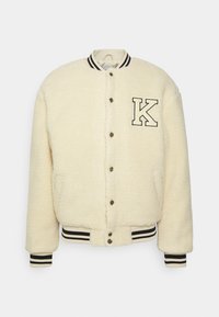 Cream-colored, fluffy faux fur jacket with a button front, ribbed cuffs, and hem. Features a patch with the letter "K" on the chest.