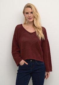 Burgundský pletený sveter s výstrihom do V, s voľným strihom, veľkým otvoreným vzorom a spustenými ramenami, skombinovaný s tmavomodrými džínsami.