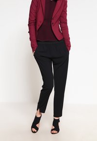 Pantalones negros ajustados con una cintura elástica, combinados con una chaqueta burdeos y una blusa a juego. Unos tacones negros de punta abierta completan el conjunto.