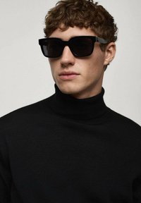 Sonnenbrille mit einem dicken schwarzen Kunststoffrahmen und dunklen Gläsern, getragen von einer Person in einem schwarzen Rollkragenpullover vor einem neutralen Hintergrund.