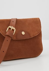 Sac bandoulière en daim marron avec bandoulière en cuir et boucle dorée, présentant un petit détail de bouton doré sur le rabat avant.