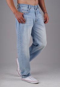 Man wearing light blue jeans and white sneakers, standing with one leg slightly bent against a plain gray background.