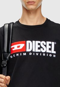 Melna kokvilnas T-krekls ar lielu balto un sarkano izšūto "DIESEL" emblēmu un tekstu "DENIM DIVISION" zem tās. Iekļauts melns siksniņš.