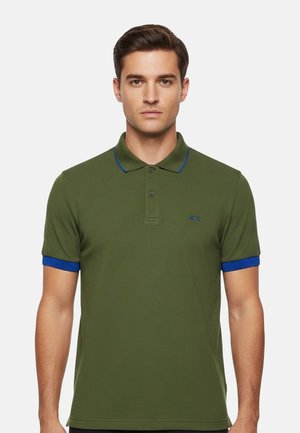 Uomo che indossa una polo verde oliva con colletto e polsini blu, in piedi davanti a uno sfondo chiaro e uniforme.