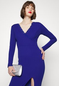 Vestido azul royal com mangas longas, decote em V profundo, com detalhes em renda e uma fenda até à coxa. Bolsa de mão prateada segurada na mão esquerda.