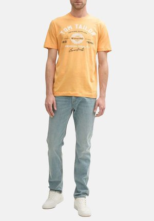 T-shirt en coton jaune avec col rond, présentant un logo de marque imprimé en blanc et du texte. Associée à un jean bleu clair et des sneakers blanches.