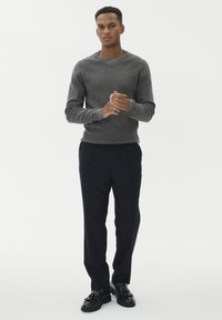 Muškarac stoji s rukama sklopljenim, odjeven u sivi pleteni pulover, crne hlače i crne kožne mokasine, nasuprot bijele pozadine.