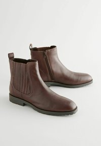 Botas de cuero marrón hasta el tobillo con paneles elásticos laterales, una lengüeta trasera y cierre de cremallera. Textura suave y suela plana de goma.