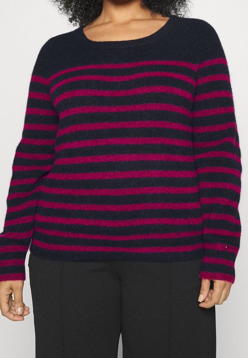 Femme portant un pull bleu marine foncé avec des rayures horizontales magenta et un pantalon noir, montrée des épaules aux hanches.