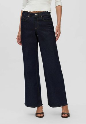 Femme portant un jean large taille haute bleu foncé, un top court blanc et des talons ouverts noirs, debout sur un fond gris.