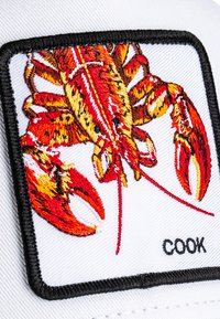 Lobster rouge et jaune brodé sur une patte blanche, bordée de couture noire, à côté du mot "COOK" en texte noir.