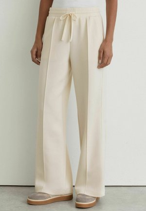 Person wearing cream wide-leg pants with drawstring waist and beige sneakers, standing against a light wall background.