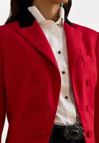 Červený dvojradový blejzer s čiernym golierom, skombinovaný s bielou košeľou s gombíkmi v čiernej farbe a dekoratívnou čiernou prackou na opasok.