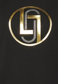 Camiseta negra con un gran logo circular dorado con un diseño estilizado de "LG" y acentos de pedrería plateada, creando una apariencia texturizada.