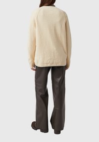 Osoba ubrana w kremowy, teksturowany sweter i ciemnobrązowe, szerokie, skórzane spodnie, stojąca tyłem na jednolitym tle.