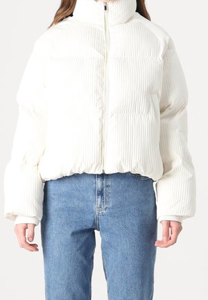 Žena v bílé prošívané bundě na zip a modrých džínách s vysokým pasem na jednobarevném pozadí.