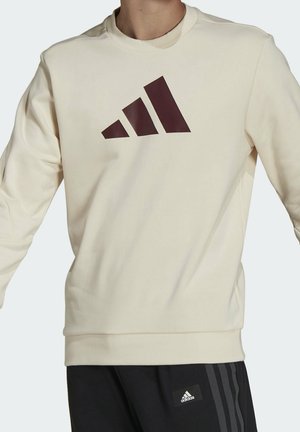 Osoba nosząca kremową bluzę z długim rękawem z dużym ciemnym logo Adidas na piersi oraz czarne spodnie Adidas z naszywką z logo i paskami.