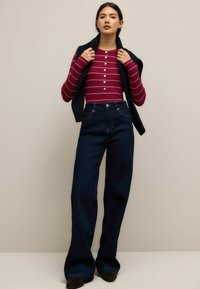 Rød og hvidstribet langærmet skjorte med knapper, parret med mørkeblå vidde jeans. Modellen holder en marineblå sweater draperet over skuldrene.