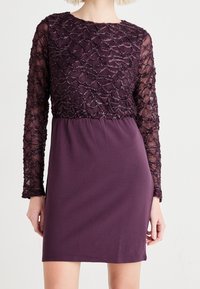 Lila klänning med en texturerad, utsmyckad överdel med långa genomskinliga ärmar och en solid, slät kjol. Pärlbroderi ger visuell intresse.