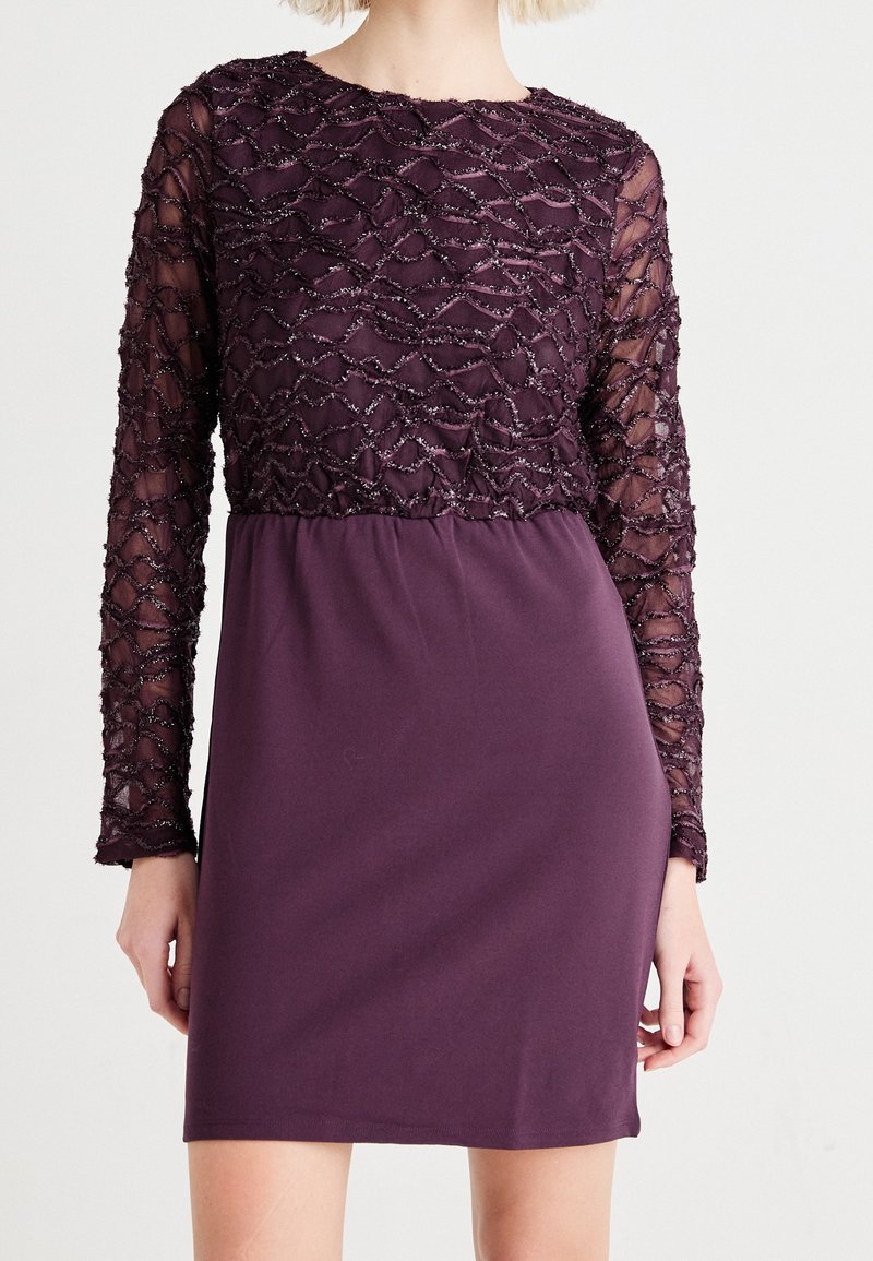 Lila klänning med en texturerad, utsmyckad överdel med långa genomskinliga ärmar och en solid, slät kjol. Pärlbroderi ger visuell intresse.