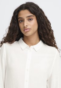 Sieviete ar gariem cirtainiem matiem valkā baltu pogājamu kreklu, skatās tieši uz priekšu ar neitrālu izteiksmi uz vienkārša fona.