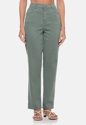 Donna che indossa pantaloni verde oliva a vita alta e gamba dritta e sandali beige con tacco e cinturini, in piedi davanti a uno sfondo bianco.