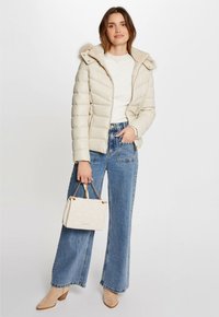 Giacca imbottita color crema con cappuccio in pelliccia sintetica, abbinata a jeans blu a vita alta, tacchi beige e una borsa bianca trapuntata.