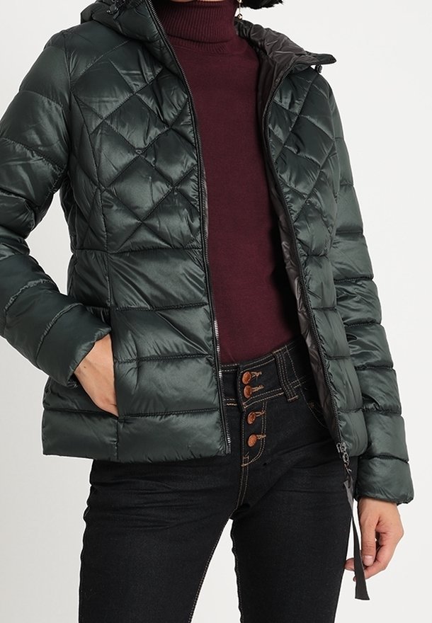 Veste noire matelassée avec fermeture éclair, portée sur un pull à col roulé bordeaux et un jean noir taille haute avec des boutons marron.