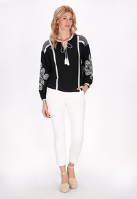 Blusa nera ricamata con disegni floreali bianchi, scollatura con lacci e pon pon, maniche lunghe, abbinata a pantaloni bianchi e scarpe chiare.