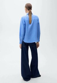 Frau mit langem Pferdeschwanz, die ein hellblaues Shirt und weite dunkelblaue Jeans trägt, steht mit dem Rücken zu einer schlichten Hintergrund.
