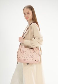 Hellrosa Stoff-Schultertasche mit floraler Stickerei, weicher Textur und strukturiertem Design. Enthält einen braunen Riemen und subtile Akzente.